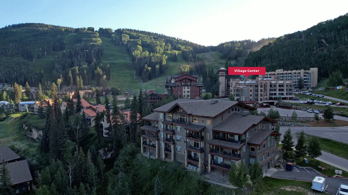 An inviting view of the resort's Village Center with residential buildings nestled against a picturesque mountain backdrop. The scene features prominent signage, open communal spaces, and a sense of community, suggesting easy access to both village events and outdoor recreation.