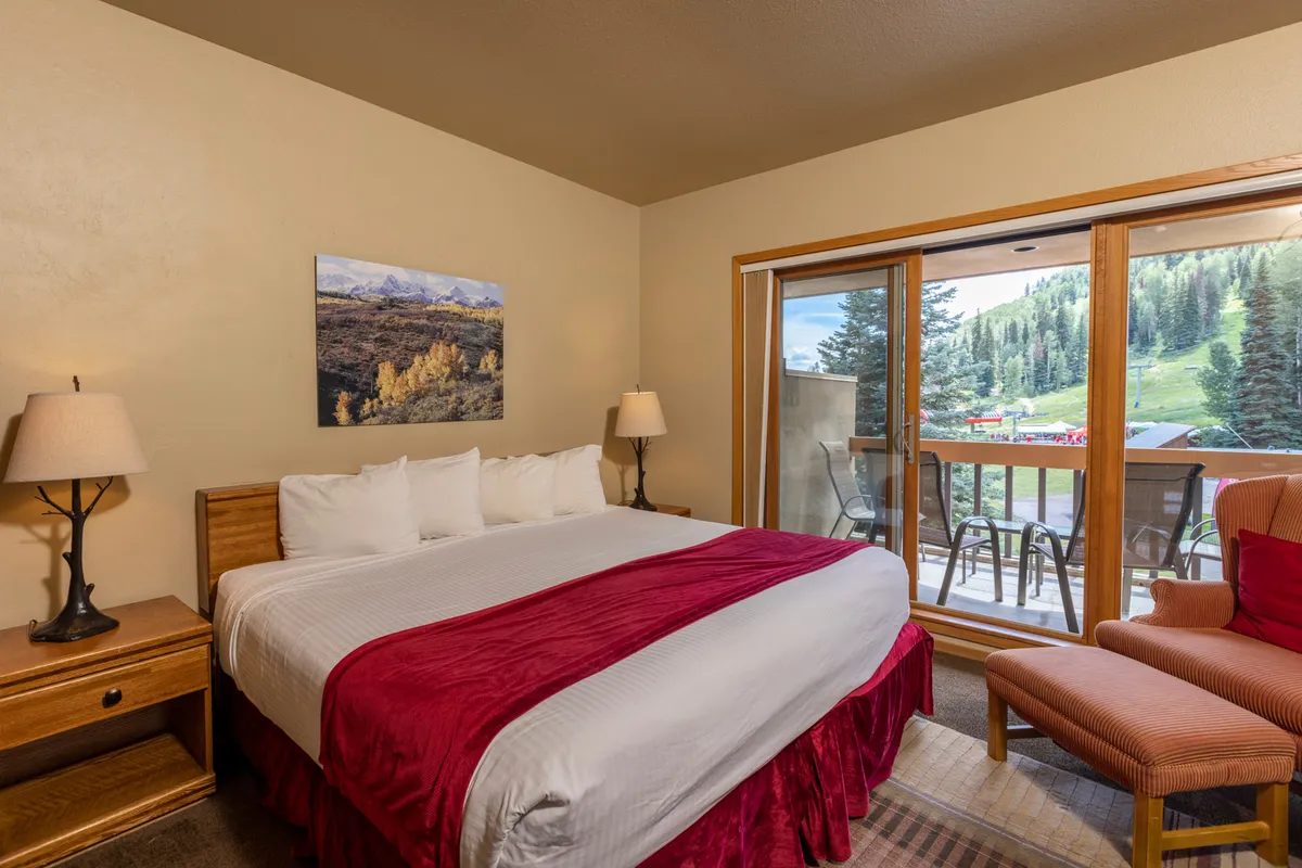 A cozy bedroom featuring a bed with red and white linens, positioned beside a large window that frames a serene mountain view. An inviting armchair sits in the corner, creating a perfect nook for relaxing with a book or sipping tea while basking in natural light.