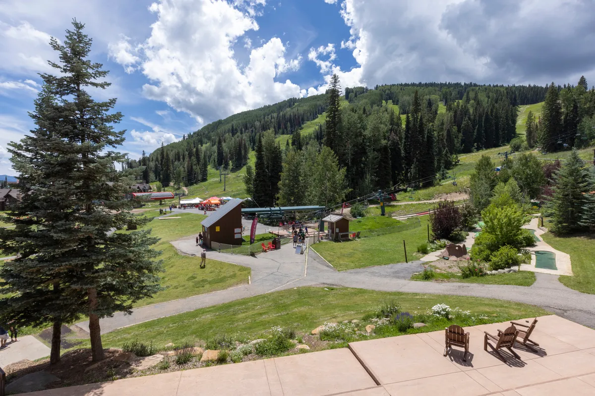 This vibrant exterior shot captures a lively mountain setting with people walking, beautiful green spaces, outdoor seating, a visible ski lift, and stunning mountain views in the background. The scene radiates activity and the sense of a bustling, family-friendly resort community surrounded by nature.