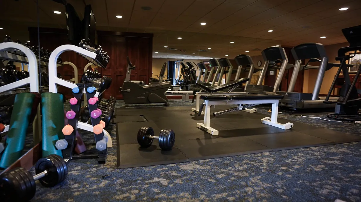 This image features a well-equipped fitness center with dumbbells, treadmills, weight bench, and exercise mats. The space is bright and designed for group and solo workouts, reflecting an active lifestyle and supporting various fitness routines.