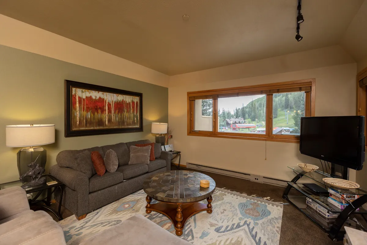 A cozy, intimate living room scene with a plush sofa, a coffee table perfect for games or snacks, artwork that adds warmth, and a window framing the sunset over the mountains. The space radiates comfort and invites both socializing and peaceful relaxation.