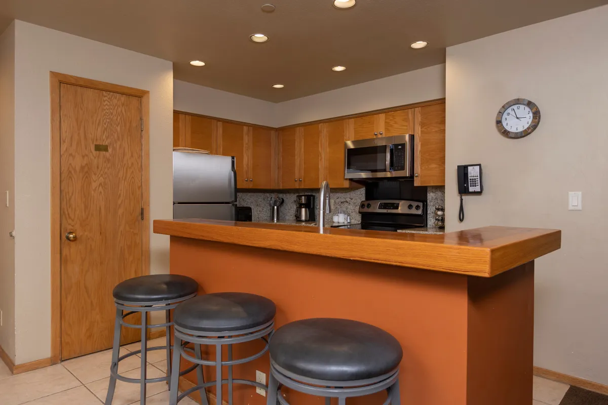 This image showcases a welcoming kitchen centered around a spacious island, with stools for casual seating. The kitchen features modern appliances, warm wood cabinetry, and a prominent clock, all bathed in natural light. The setup is perfect for both cooking and socializing, reflecting the open-concept design and inviting atmosphere described in the section.
