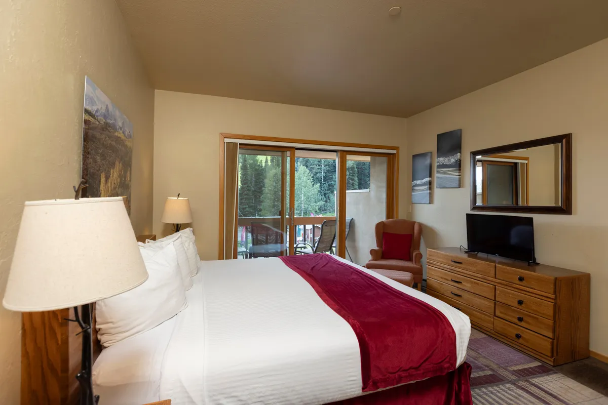 A warm bedroom scene showing a comfortable bed, a balcony with a view, an armchair, a chest of drawers, and a gentle glow from a lamp. The scene evokes relaxation—whether for morning coffee on the balcony or quiet evening conversations inside.