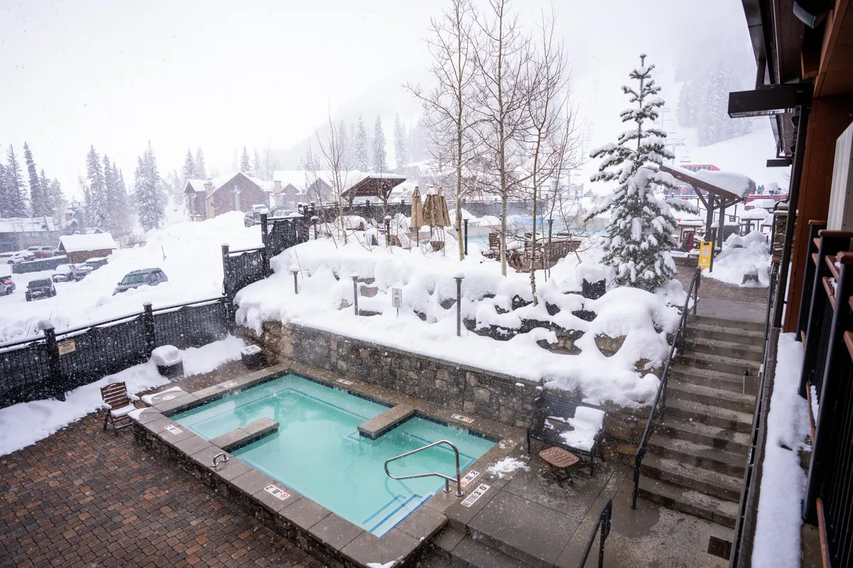 An inviting outdoor hot tub surrounded by a snow-covered landscape, with steam rising as guests soak and enjoy the tranquil wintry atmosphere. The stone steps and nearby trees add a sense of privacy and natural beauty, making the space feel both exclusive and relaxing.