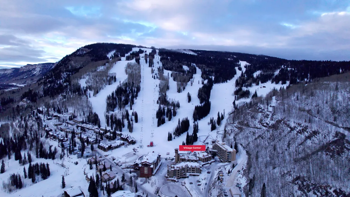 A sweeping exterior view of the Village Center set against snow-dusted ski slopes and lush surrounding forests. The image captures both the immediate access to the mountain and the inviting ambiance of the resort community.