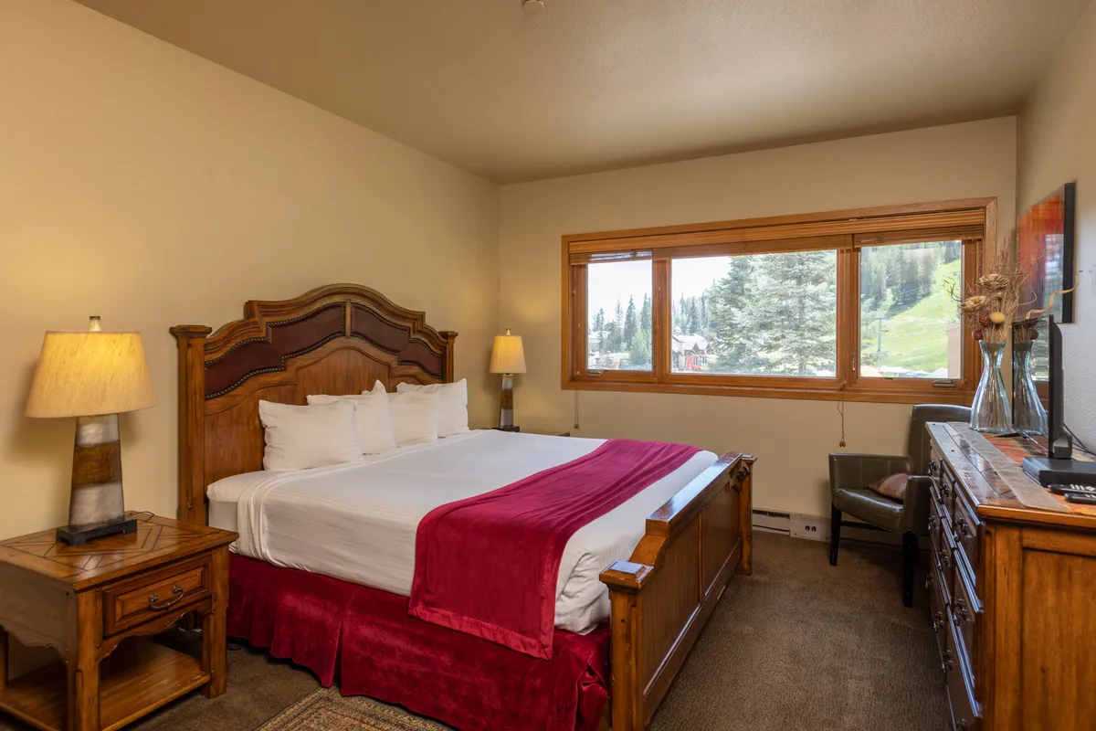 A spacious bedroom with a king-size bed adorned with a decorative headboard. The room is brightened by a large window offering a scenic mountain vista. A wooden dresser and a nightstand with a lamp add comfort and utility, highlighting a restful space for unwinding after the day’s adventures.