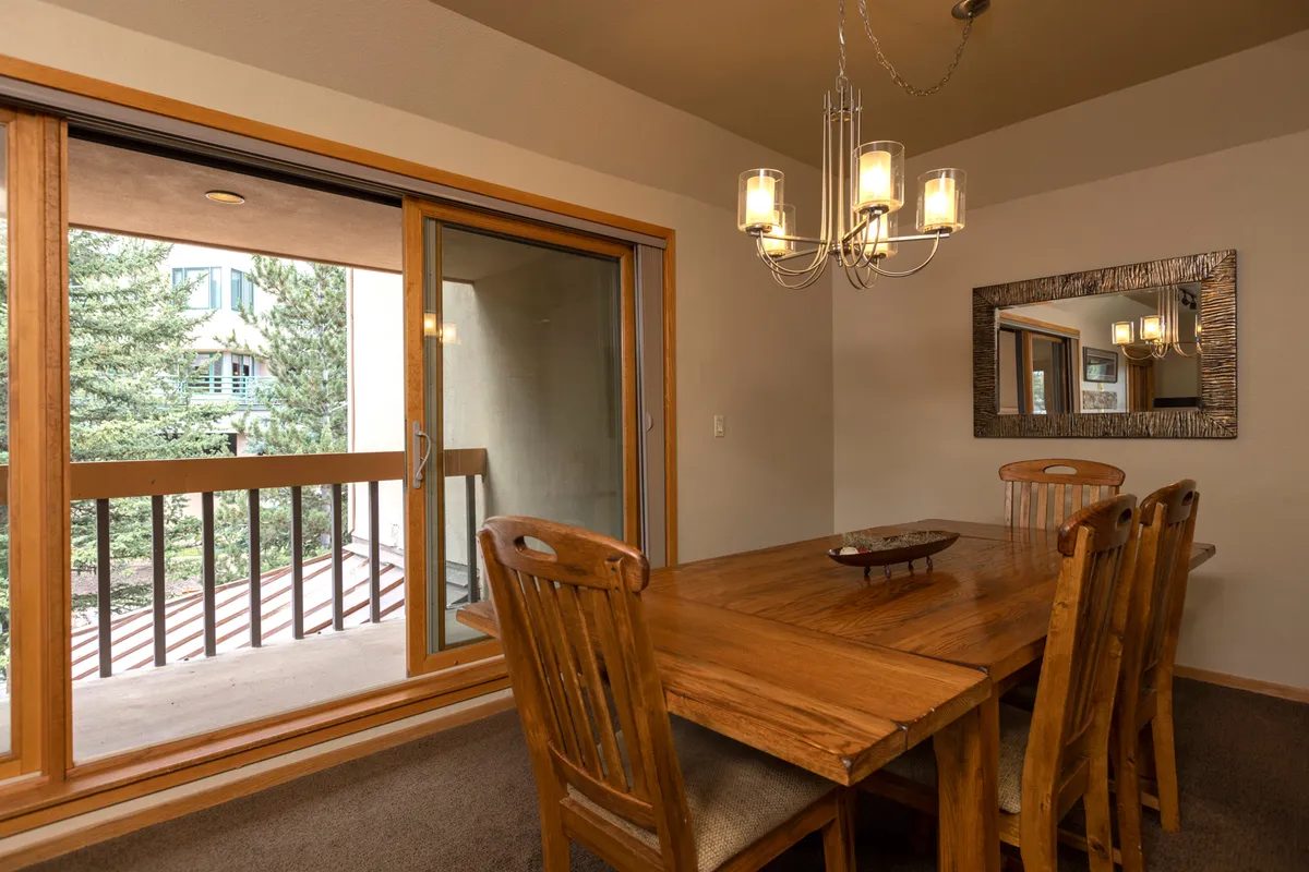 A warm and inviting dining area is depicted, featuring a large wooden table beneath a stylish chandelier. Natural light streams in through a large window, while a mirror adds depth to the space. The setup encourages group meals, celebrations, and cozy conversations.