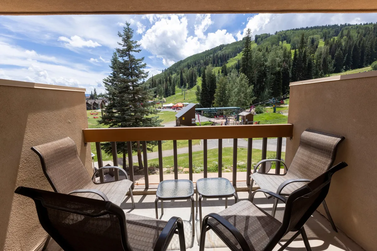 The image showcases a spacious balcony with comfortable seating, perched above a scenic mountain landscape. Towering trees and the expansive Needles Mountain Range form a breathtaking backdrop, with a clear sky overhead. The setting suggests relaxation, enjoying the panoramic views, and socializing while surrounded by nature's beauty.
