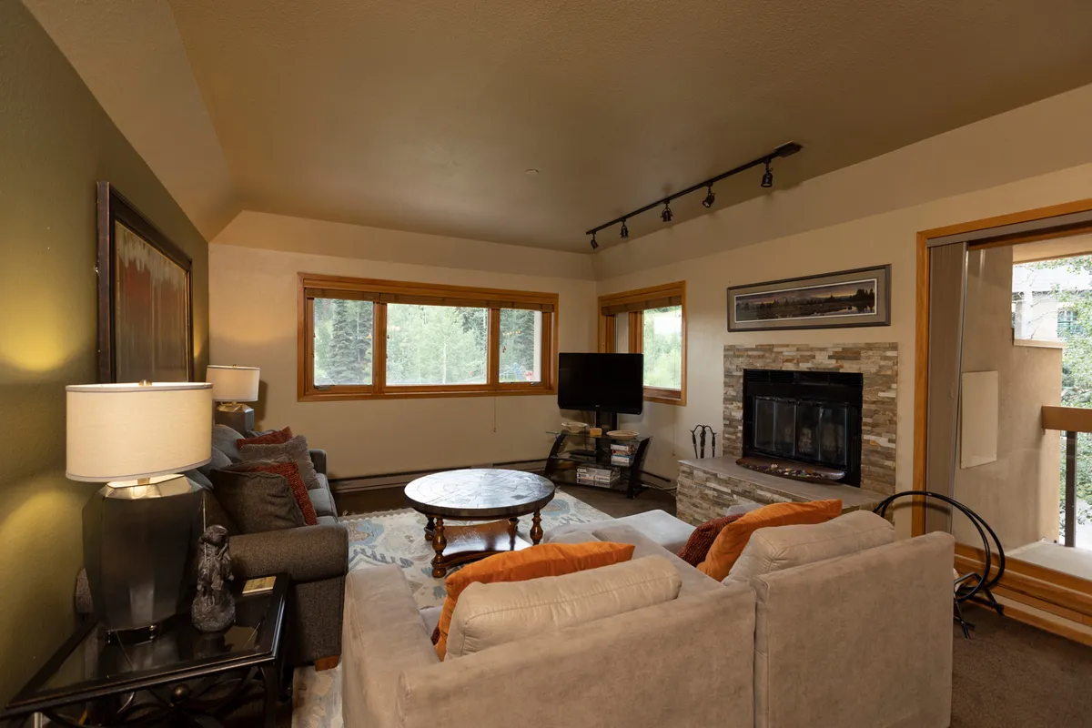 A warm and welcoming living room centered around a fireplace, with plush seating arranged to face expansive windows that fill the space with natural light and offer breathtaking mountain views. The coffee table is positioned as a gathering spot, and the overall ambiance invites relaxation and connection.